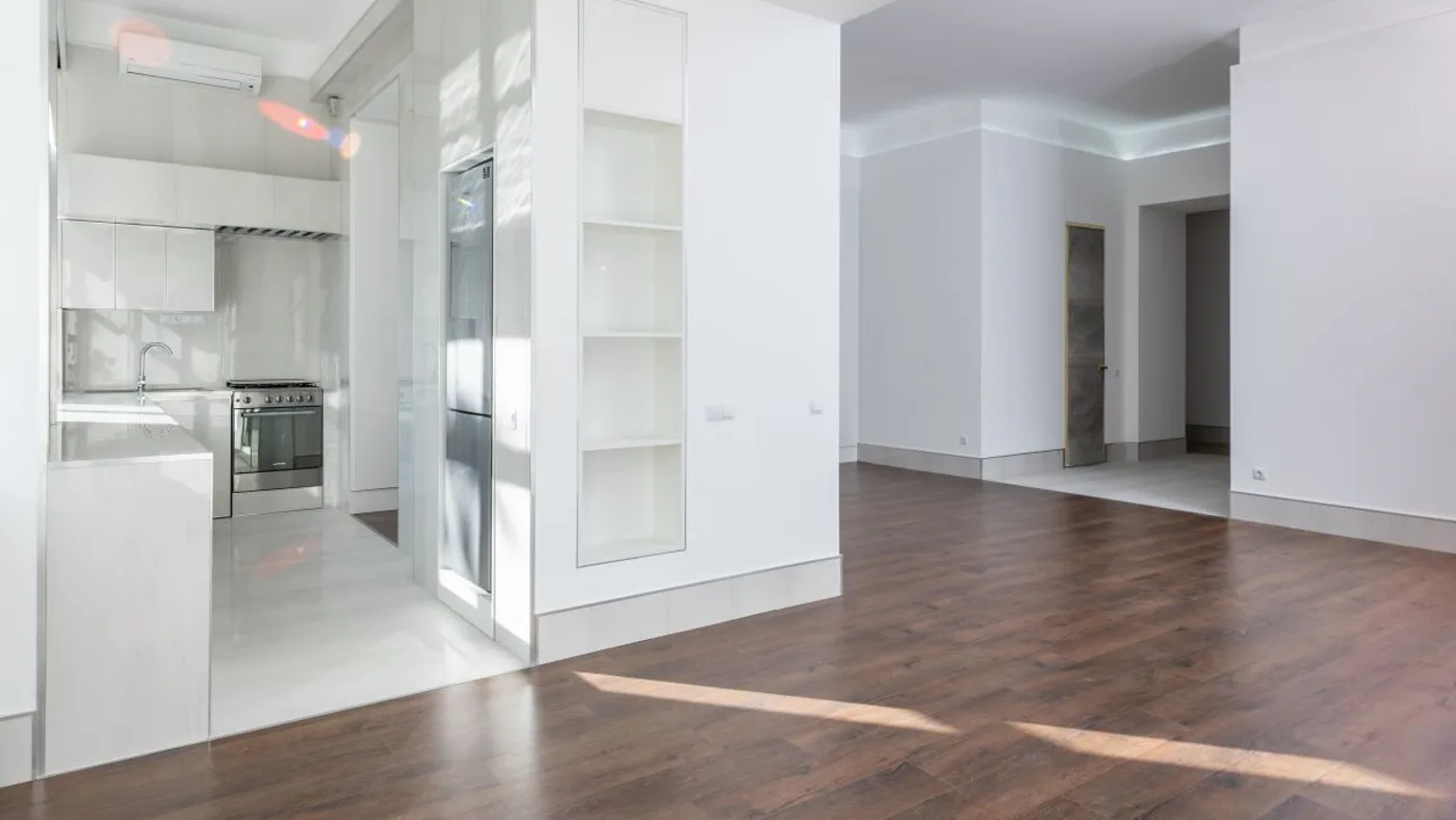 Bright emptied apartment with a fitted white kitchen, wall-mounted air conditioning unit and dark wooden flooring