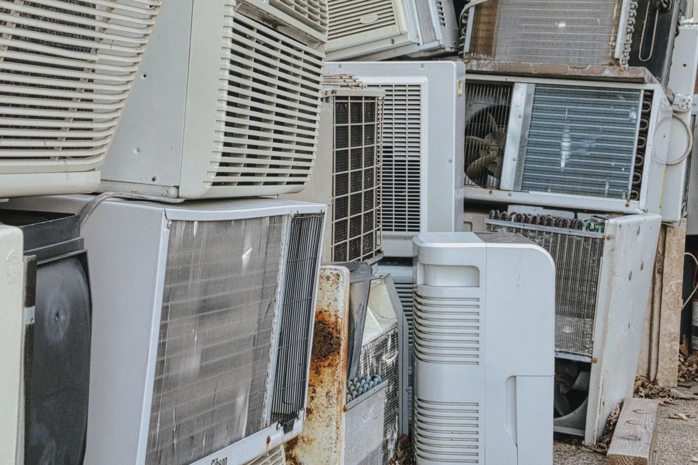 Large stack of old window and split air conditioning units piled outdoors, several showing rust and missing grilles