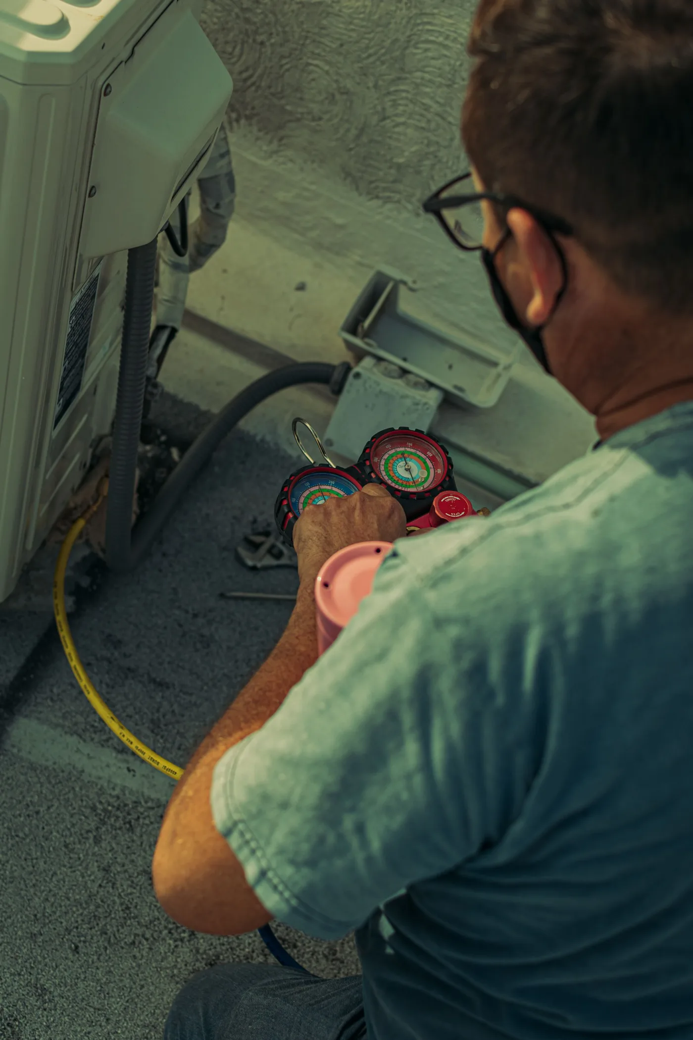 Technician kneeling beside an outdoor air conditioning unit, holding a red and blue manifold gauge set connected by hoses