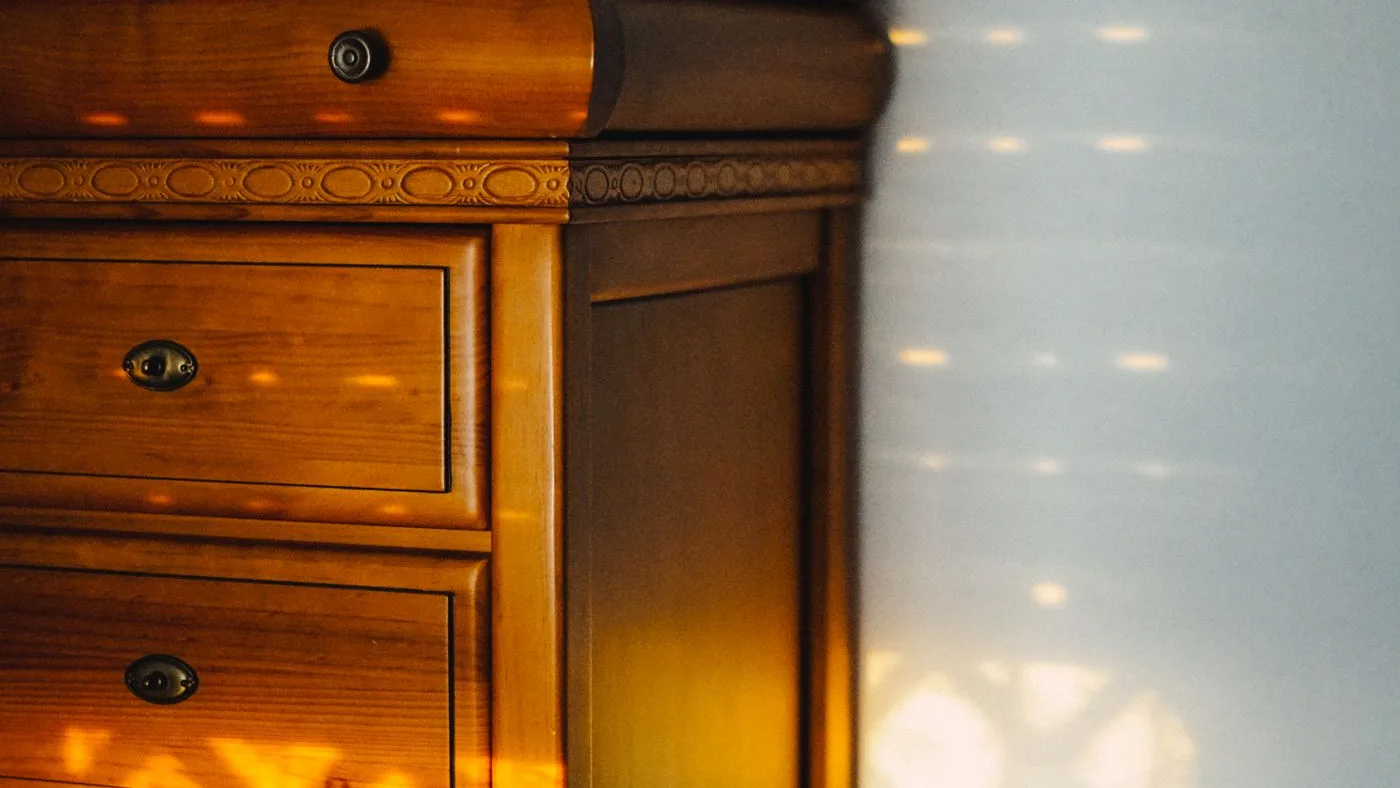 Warm-toned antique wooden chest of drawers with brass keyhole plates and a carved cornice standing beside a bright window