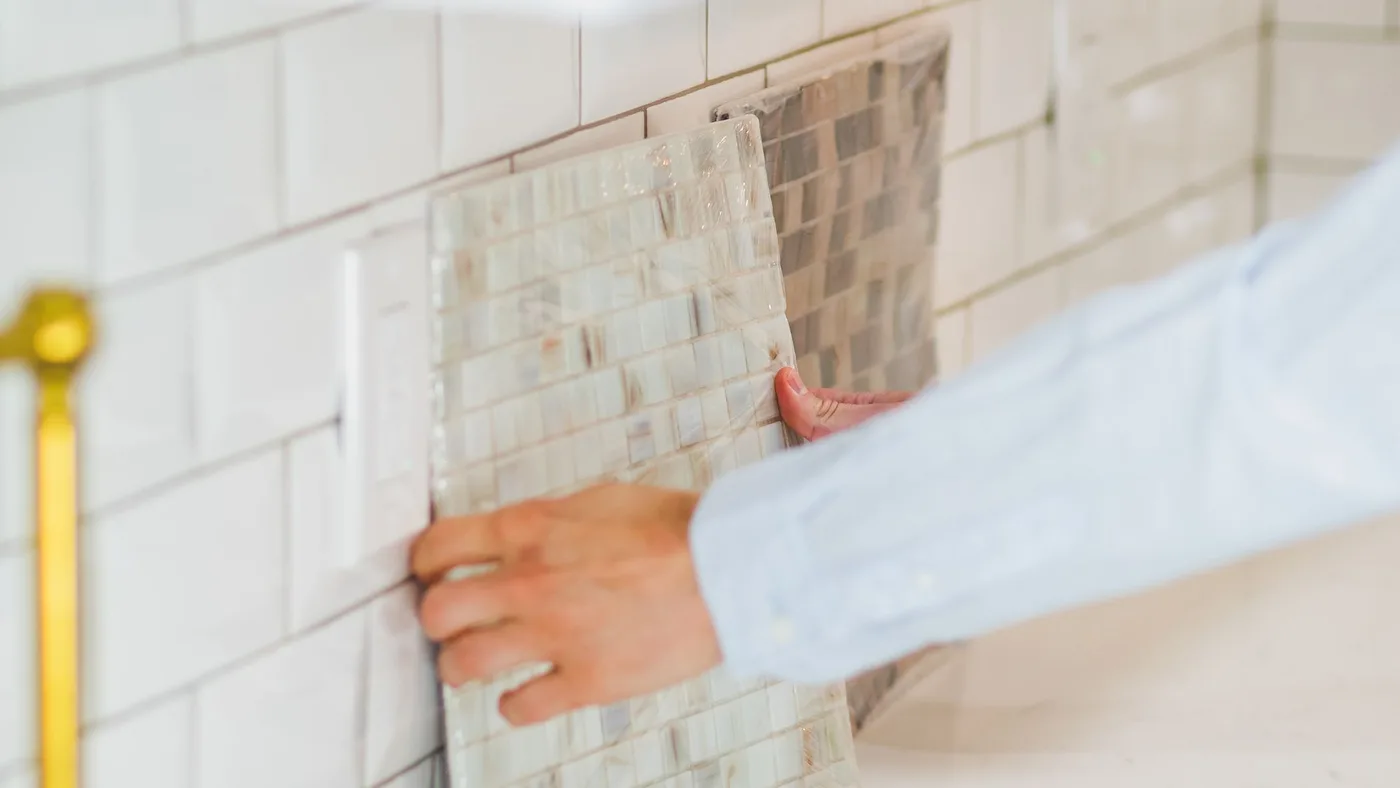 Hand pressing a mosaic tile backsplash panel into place above a kitchen counter during a renovation, with a brass tap visible in the foreground