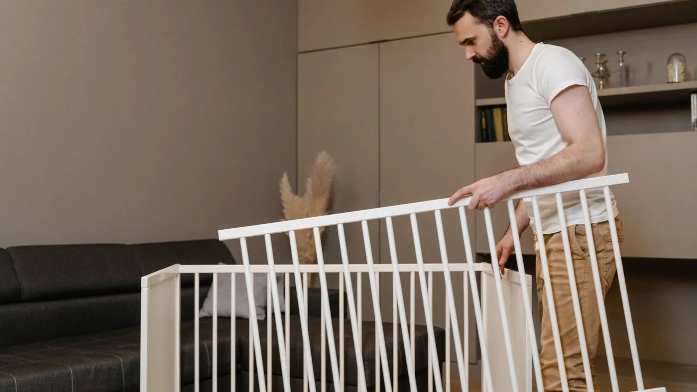 Man in a white t-shirt lifting a detached white wooden cot side panel of vertical slats away from a partly dismantled frame