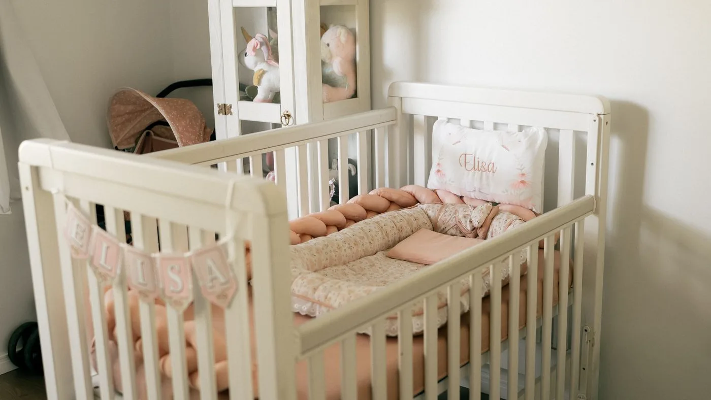 White wooden cot on castors made up with pink bedding, beside a glazed display cabinet of soft toys and a folded pram in the corner