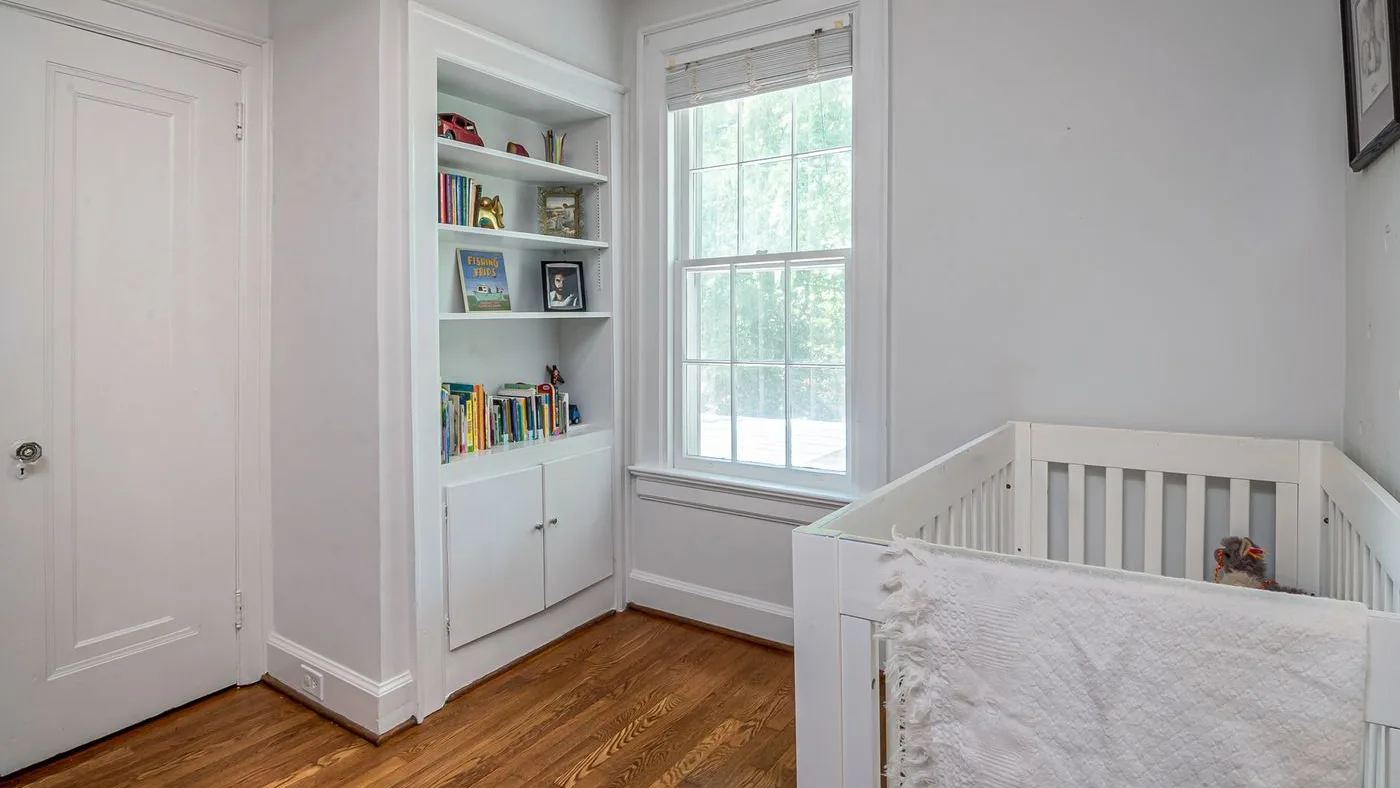 Small nursery with a white cot draped in a cream blanket beside a sash window and built-in shelves of children's books