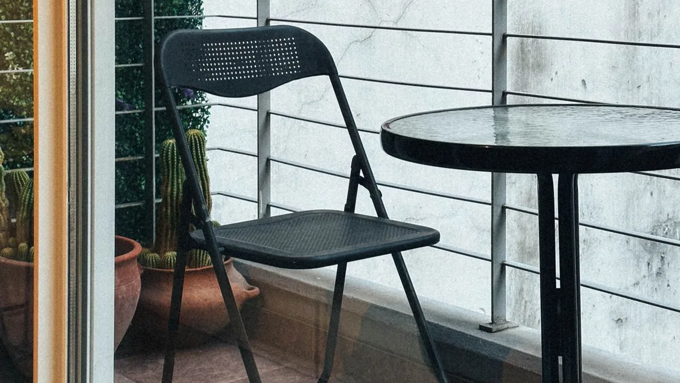 Folding metal chair and a round glass-topped table on a narrow apartment balcony with a cable railing and a potted plant in the corner