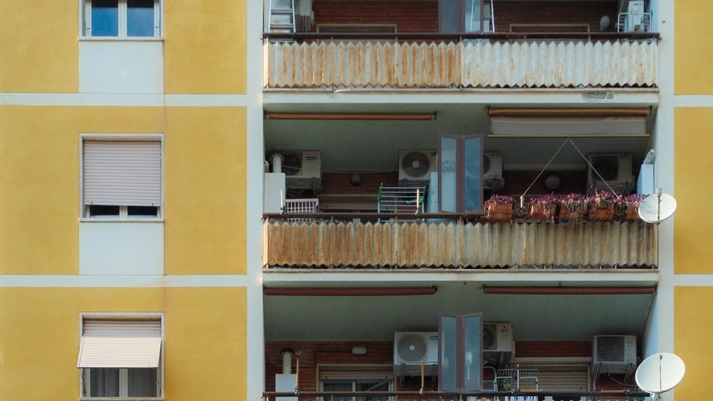 Facade of a residential apartment block showing several floors of balconies holding split air conditioning units, drying racks, satellite dishes and potted plants