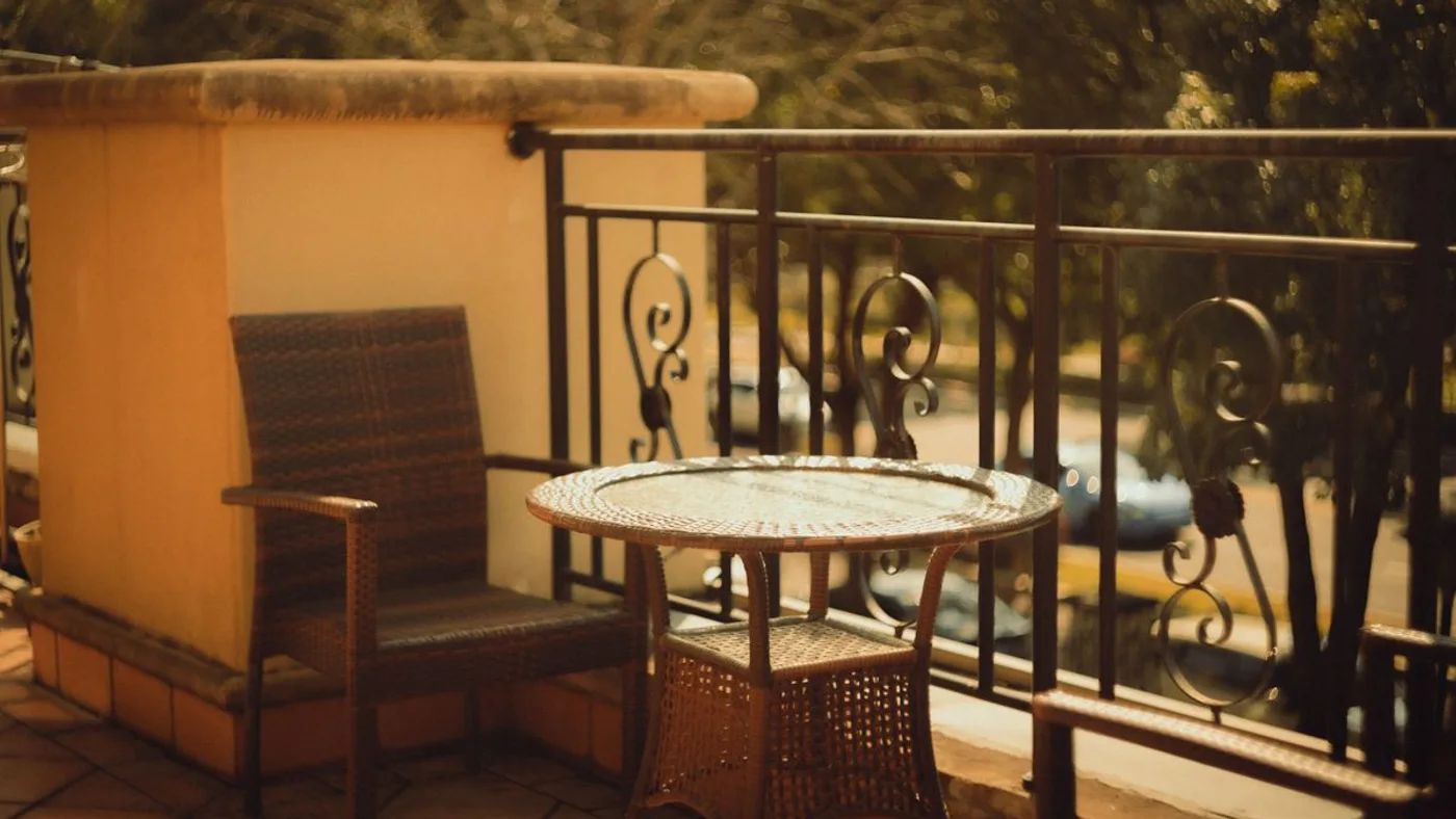 Sun-bleached balcony with a woven rattan armchair and a mosaic-topped metal table behind an ornate iron railing