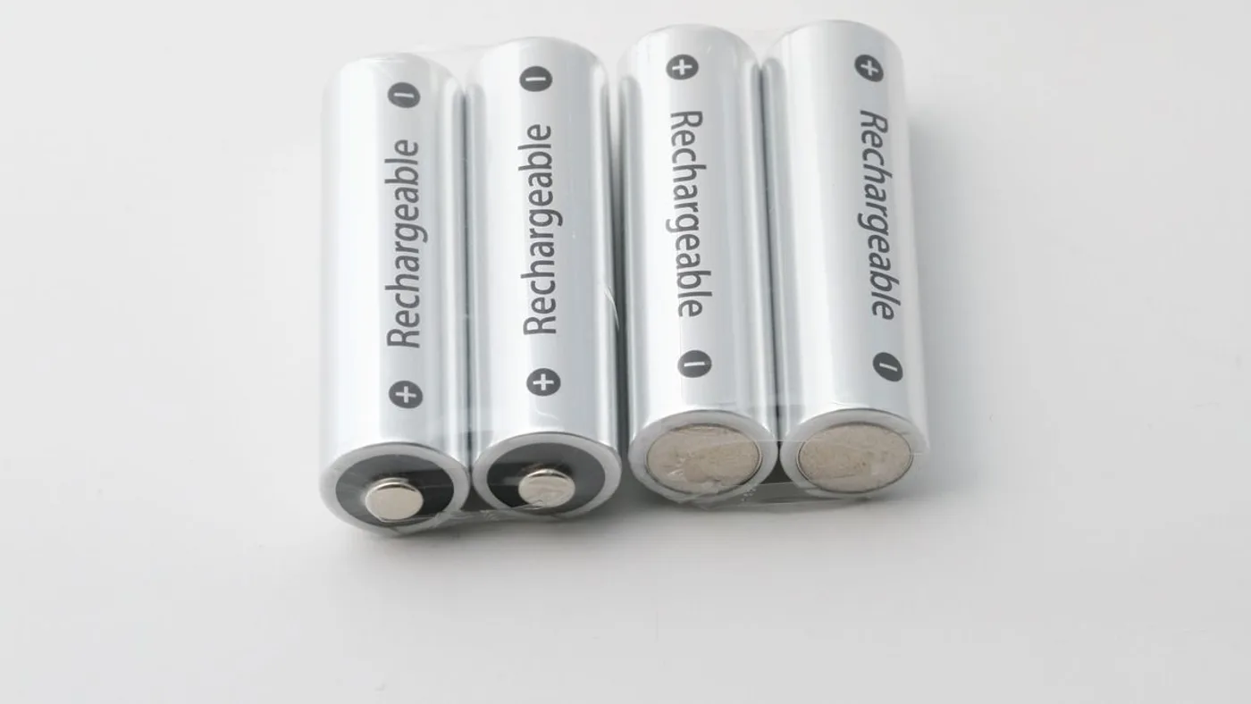 Four white rechargeable AA batteries in shrink wrap lying beside a white plug-in charger on a pale surface