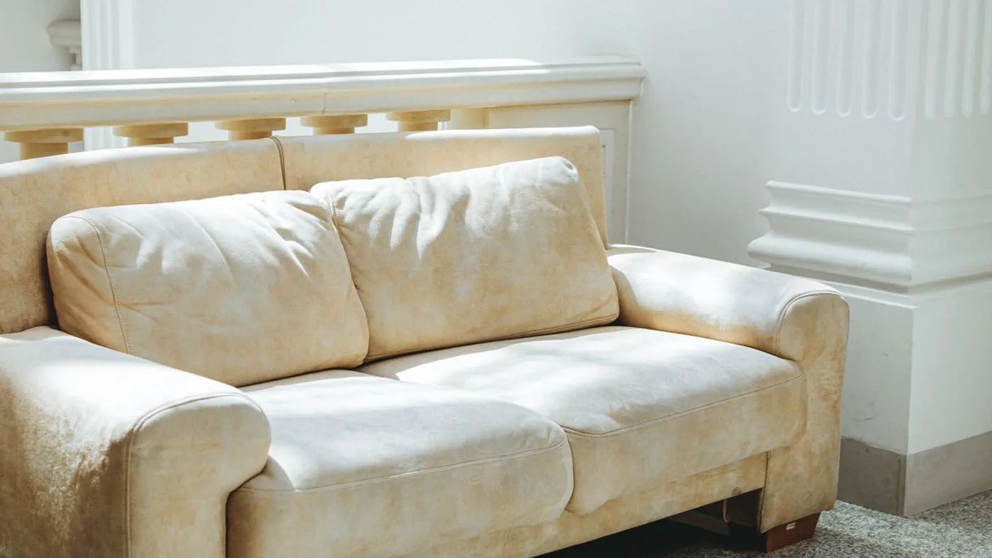 Cream upholstered two-seat sofa with worn, sun-faded cushions standing in a bright room beside a white column
