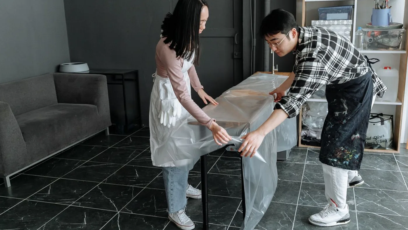 Two people in aprons and gloves wrapping a wooden table in clear plastic sheeting inside a room before moving it