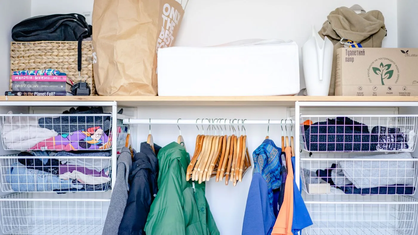 Open storage unit with coats and shirts on wooden hangers, wire baskets of folded clothing, paper bags and boxes on the shelf above