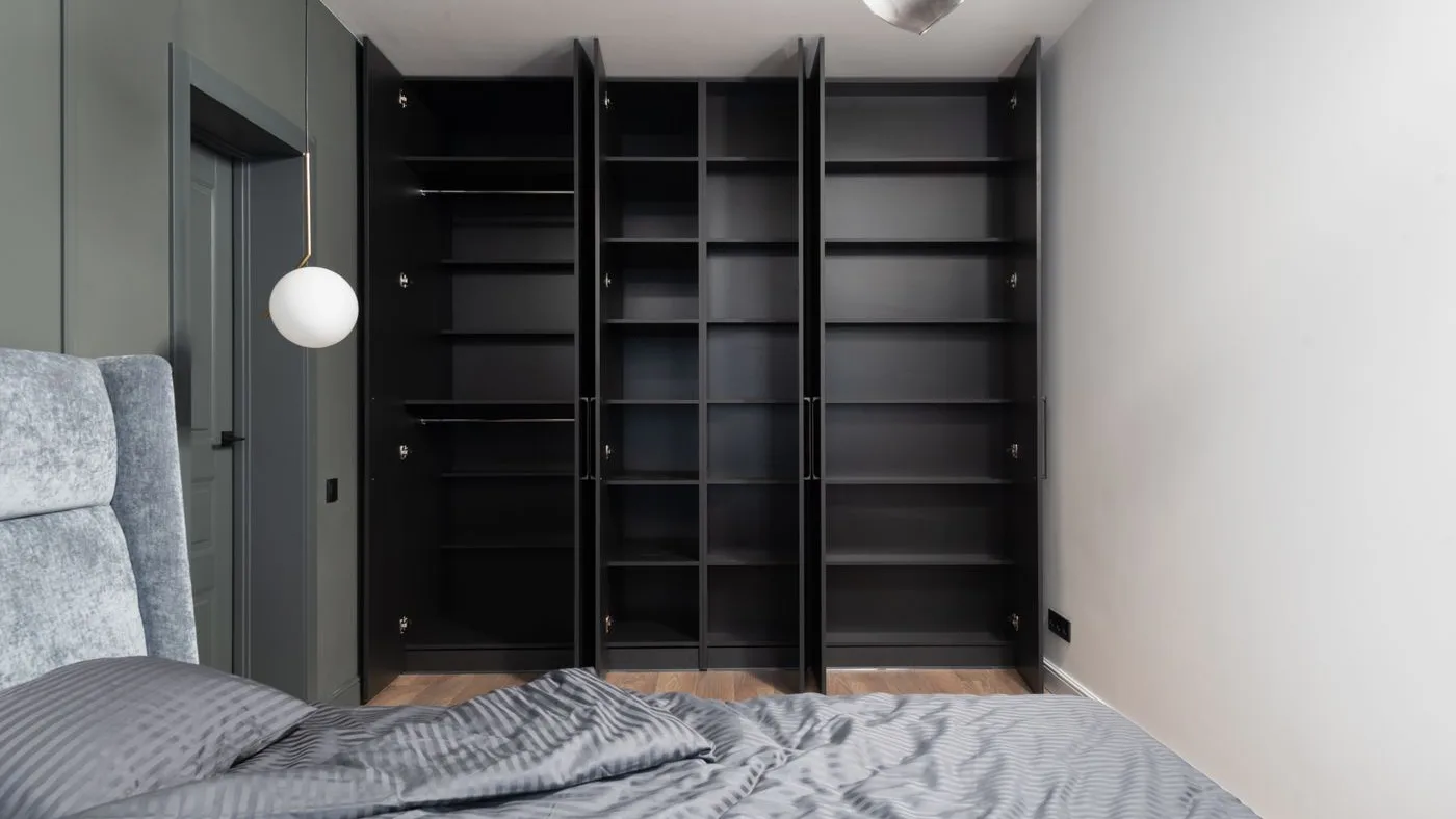 Bedroom with tall black wardrobes standing open and completely emptied of shelves and clothes beside an unmade grey striped bed