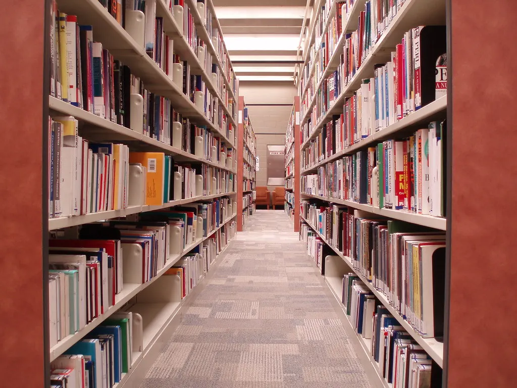 Narrow aisle between two tall library shelving units packed floor to head height with books, leading to a seating area at the end