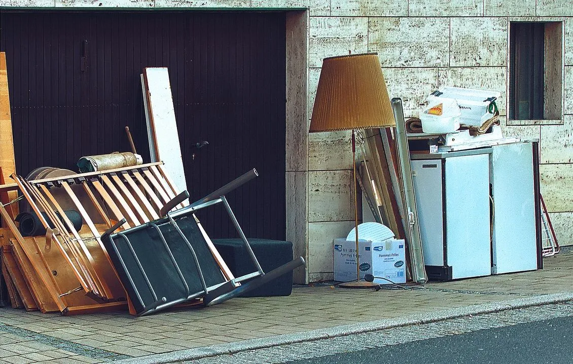 Kerbside pile of broken furniture beside a garage door, including a dismantled slatted bed base, a collapsed folding chair, loose timber panels and a floor lamp