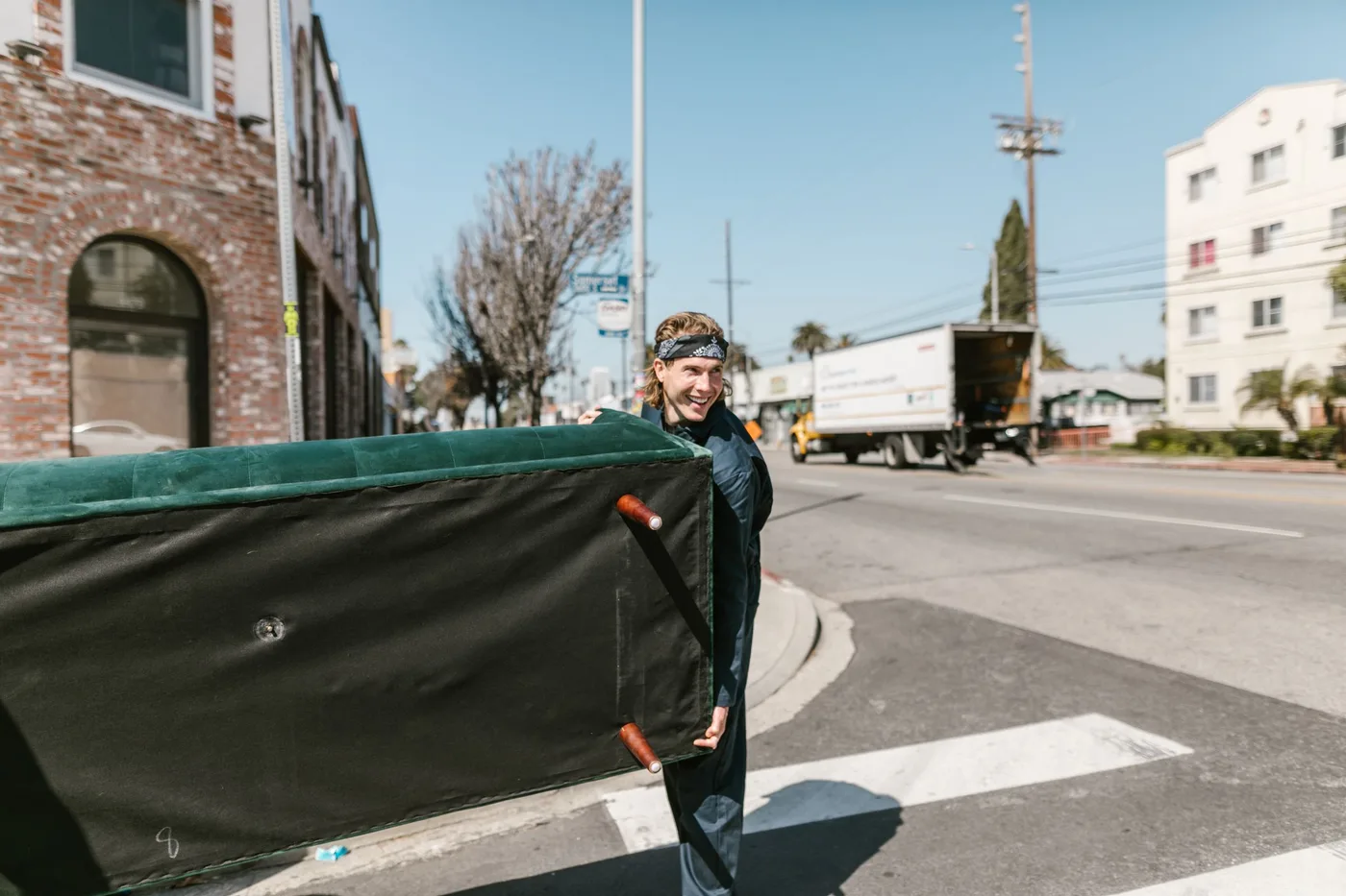 Man carrying a large green sofa across a street crossing, with a box truck parked in the background