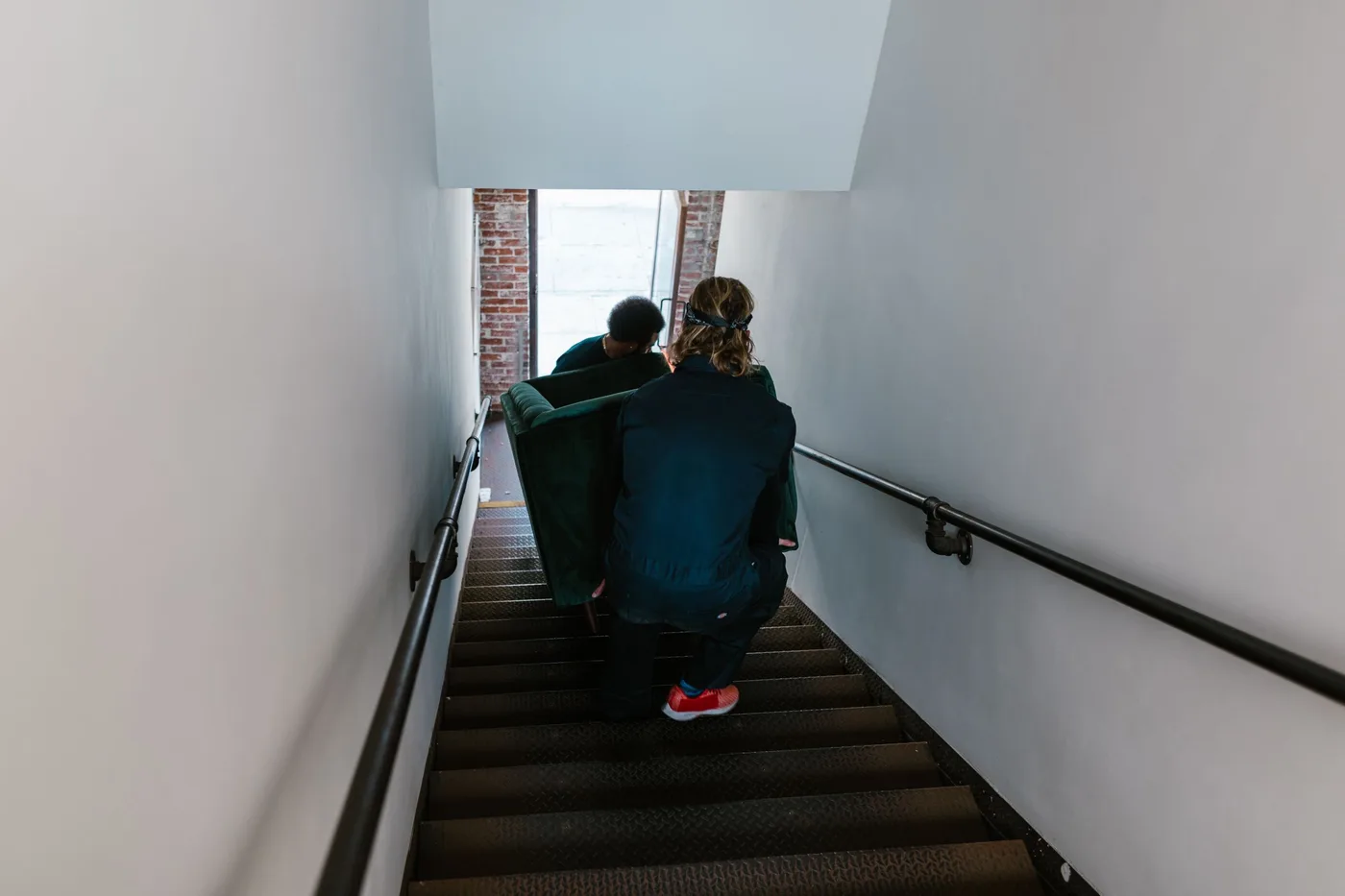 Two people in dark uniforms carrying a green sofa down a narrow indoor staircase
