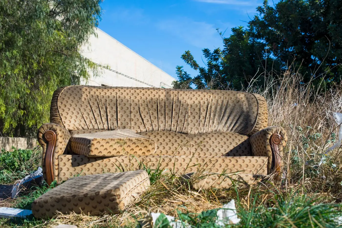 Patterned brown and gold sofa abandoned on rough ground beside a boundary wall with a cushion fallen in front of it