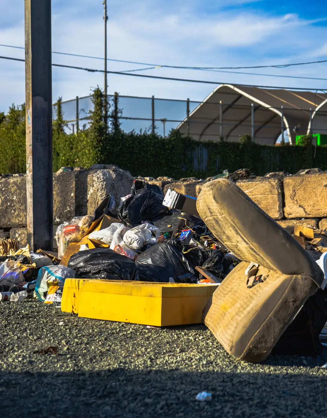 Discarded foam mattress leaning against a low wall beside a pile of black rubbish bags and scattered litter at a kerbside