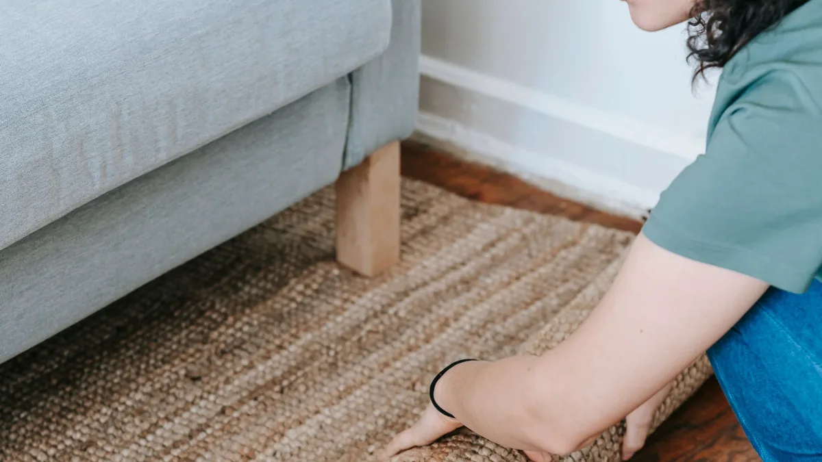 Person kneeling on a wooden floor rolling up a woven jute area rug beside a grey sofa