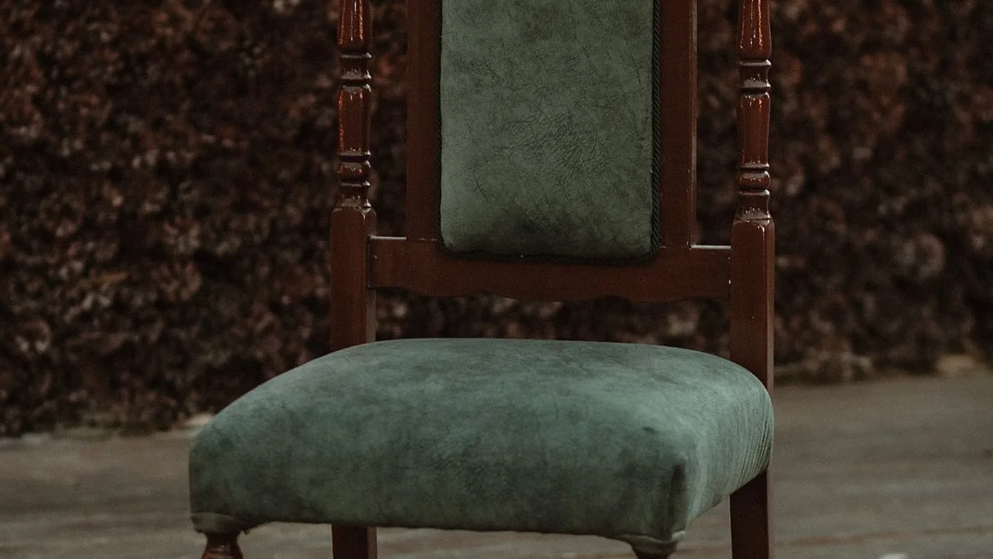 Vintage wooden chair with a green upholstered seat and backrest standing against a textured wood-panelled wall