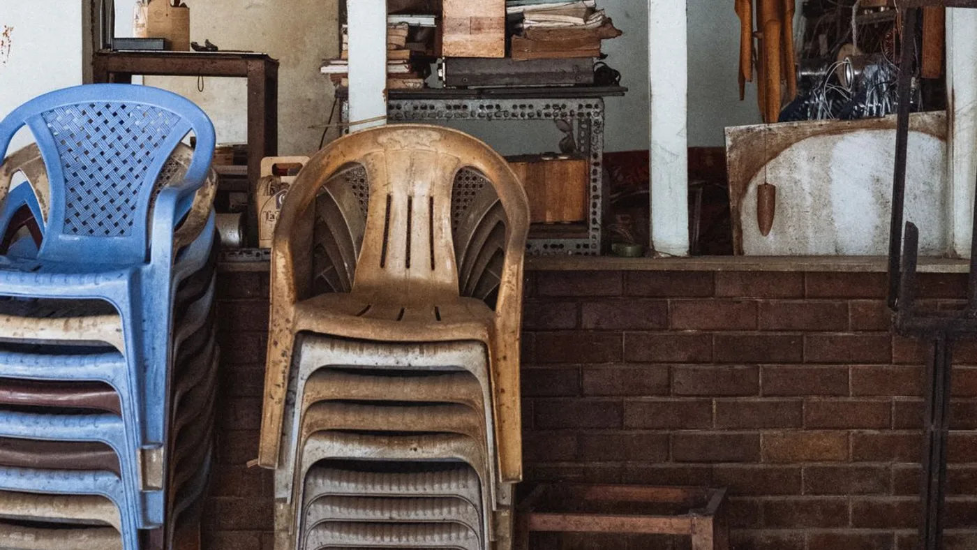 Blue and tan plastic stacking chairs piled against a brick wall in a cluttered storage area with shelving in the background