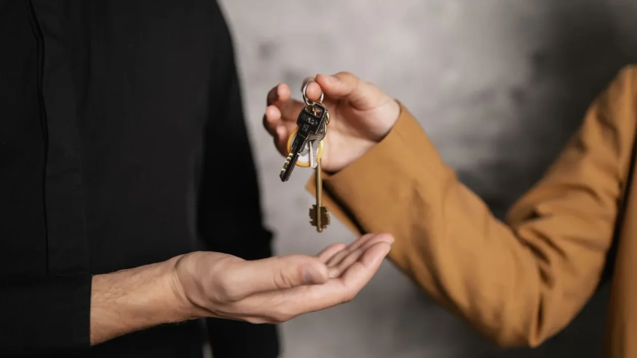 One person dropping a bunch of house keys into another person's open palm against a grey wall