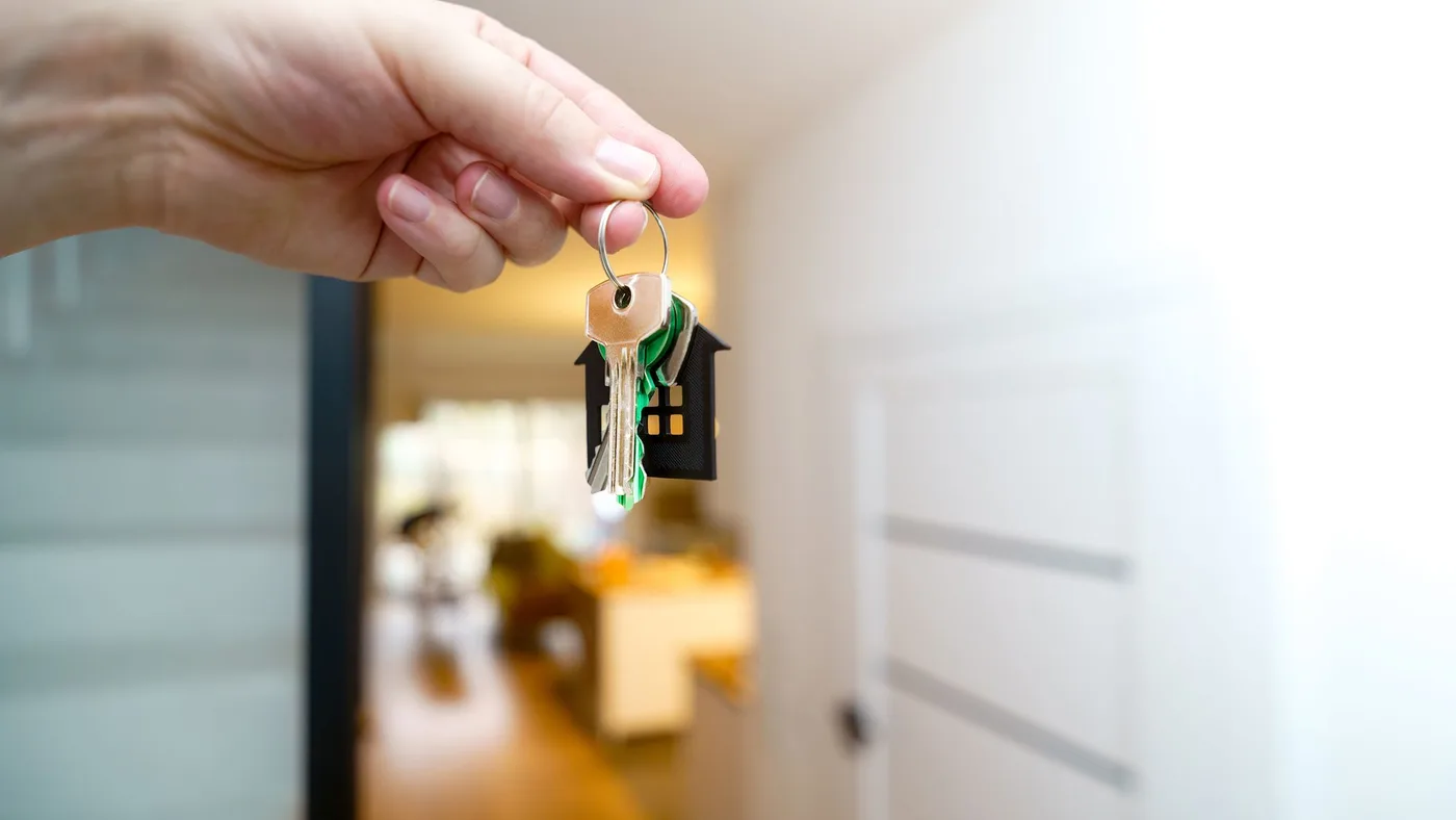 Hand holding a door key on a ring with a small house-shaped keyring, with a bright hallway and open doorway blurred behind