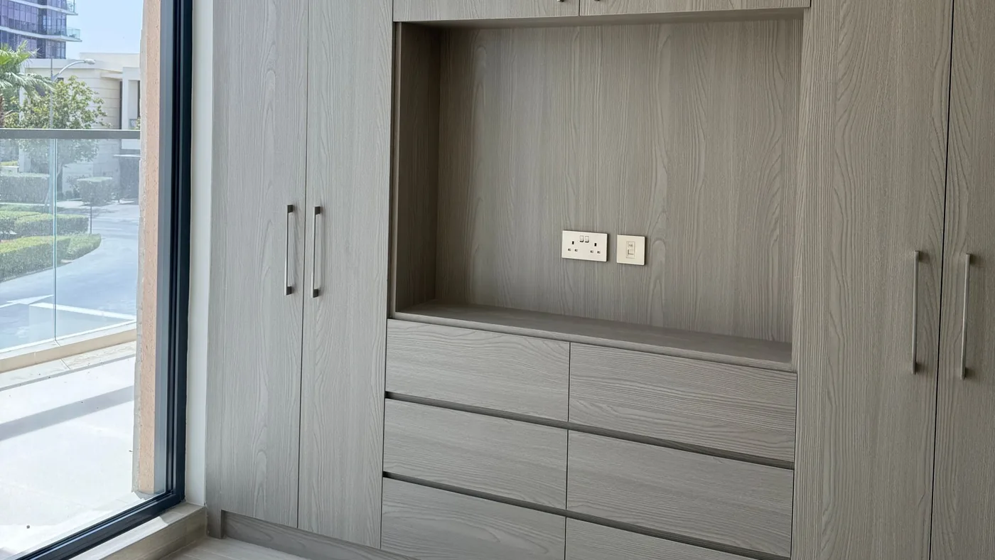 Floor-to-ceiling fitted wardrobes in pale grey wood with a central open recess and drawer bank, in an empty tiled bedroom with a tall window