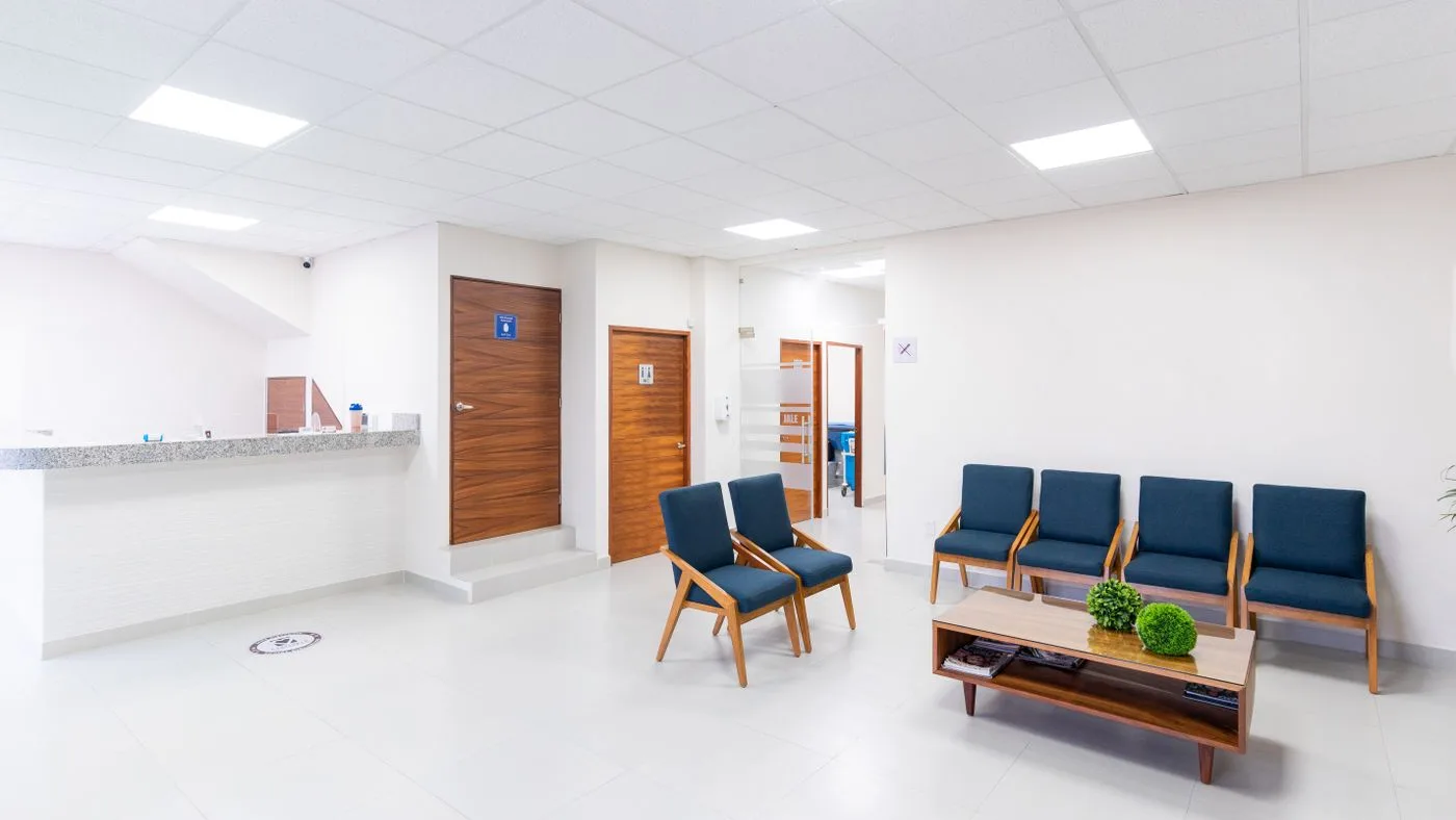 Bright clinic waiting area with a granite-topped reception counter and rows of navy upholstered wooden-framed chairs beside a low coffee table