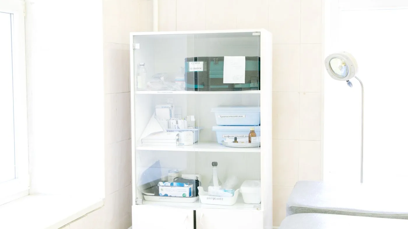 Glass-fronted white medical storage cabinet holding labelled trays, bottles and boxed supplies beside an examination couch and an adjustable lamp