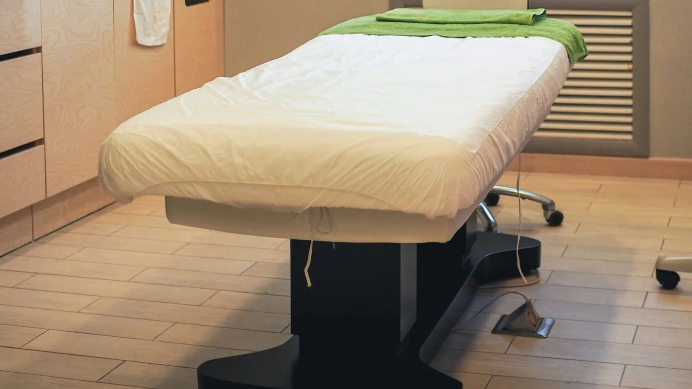 Powered treatment couch with a cream padded top and black pedestal base in a consultation room, its foot control cable trailing across the tiled floor