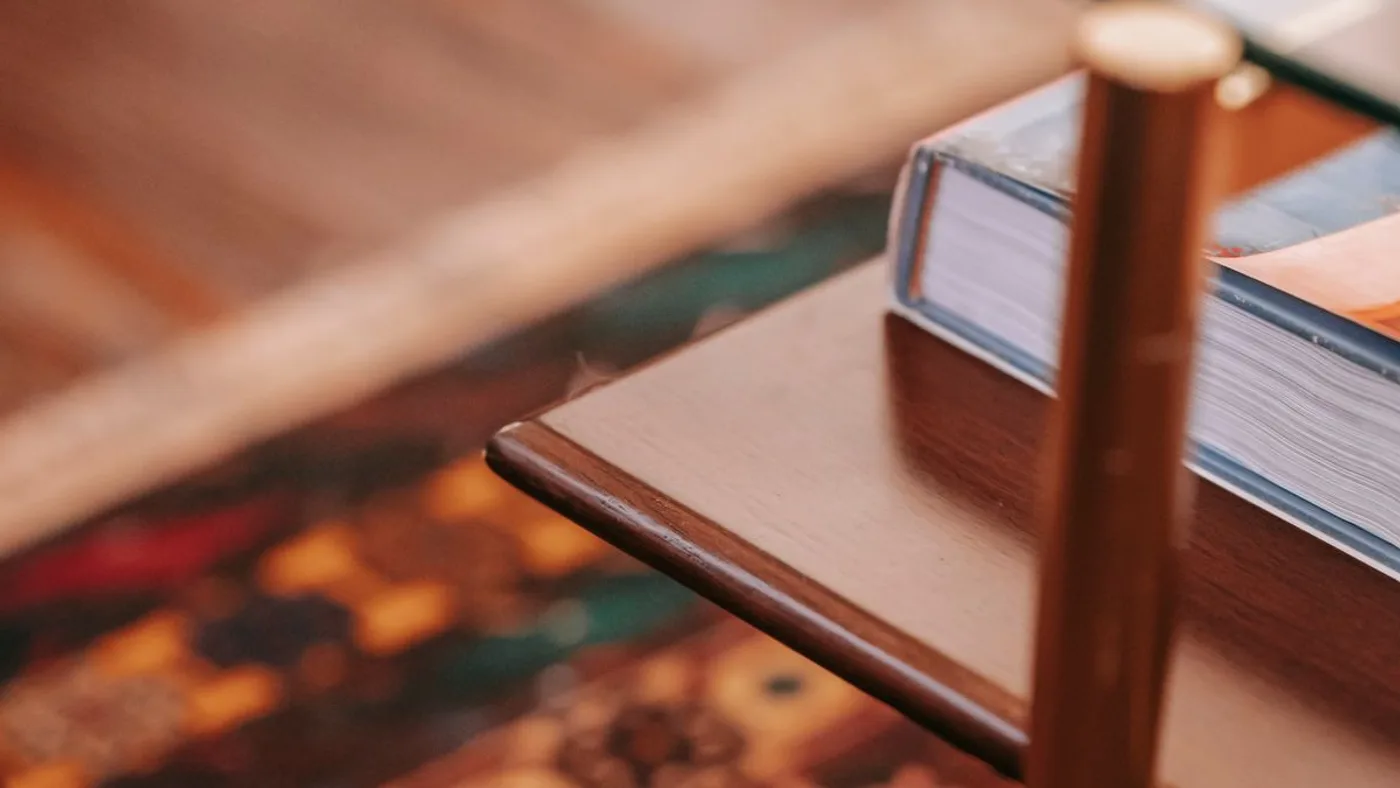 Close-up of a glass coffee table corner resting on a wooden shelf with a brass leg beside a hardback book on a patterned rug