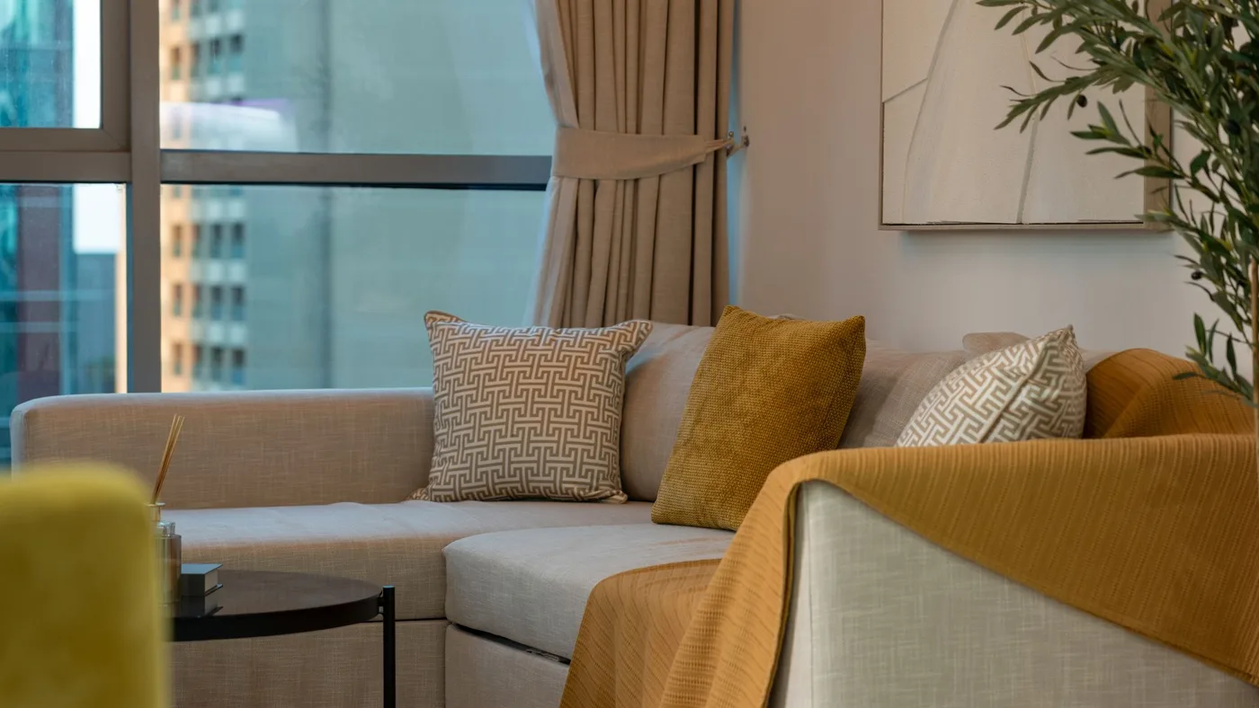 Corner of a furnished apartment living room with a beige sofa, patterned cushions and a mustard throw, with a high-rise tower visible through the window