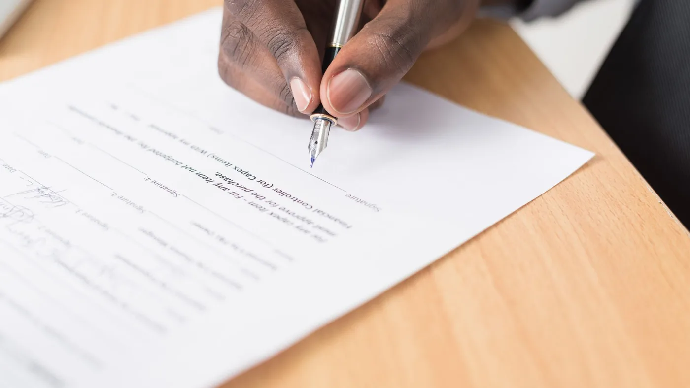Hand signing a printed purchase approval form with a fountain pen on a light wooden desk, with signature lines visible below