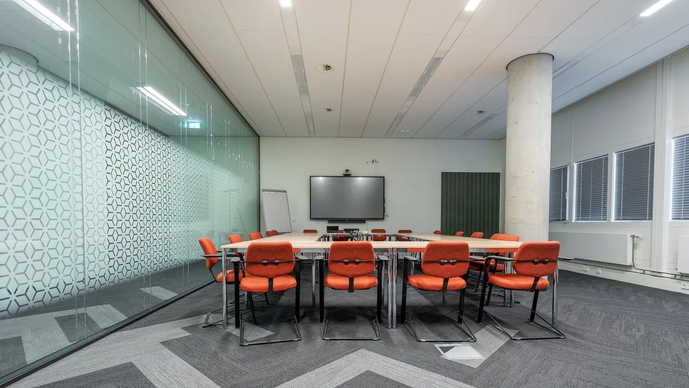Meeting room with a pale segmented conference table surrounded by orange upholstered chairs, a glass partition wall and a concrete column