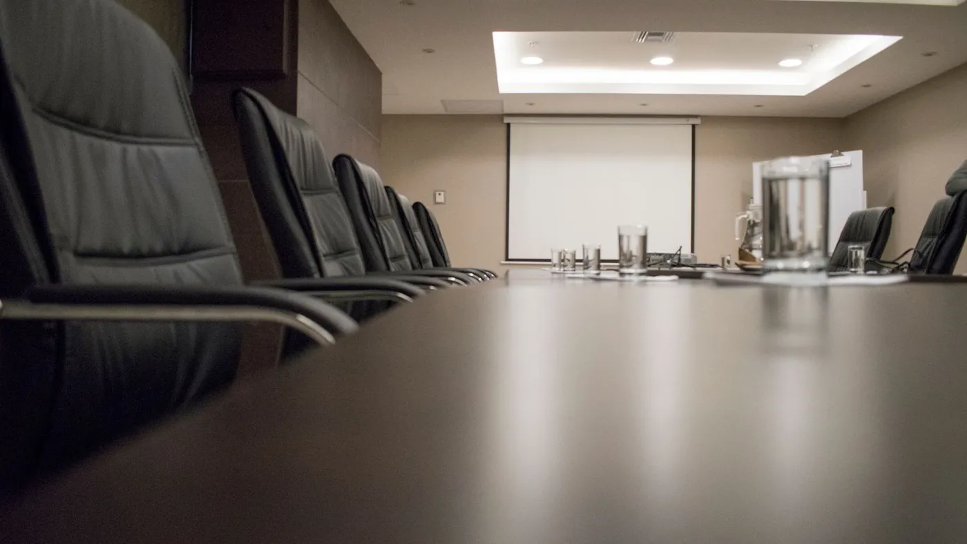 Long dark polished boardroom table photographed down its length with a row of black leather executive chairs and a projector screen at the far end