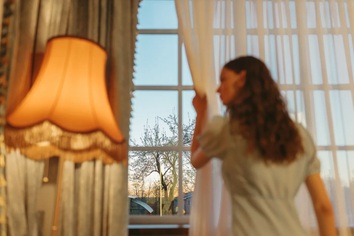 Person standing at a tall window holding a sheer white curtain aside, with a heavier drape and a fringed lamp in the foreground