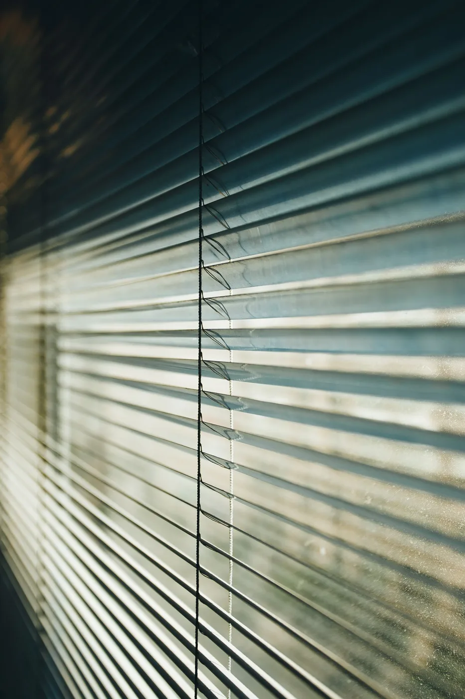 Close-up of horizontal aluminium venetian blind slats with a pull cord, backlit by sunlight and showing a film of dust