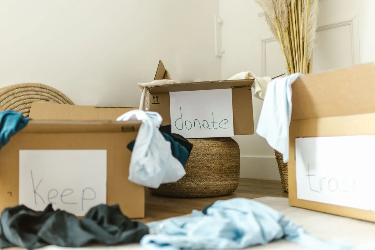 Three cardboard boxes labelled Keep, Donate and Trash on a bedroom floor beside a woven basket and folded clothing