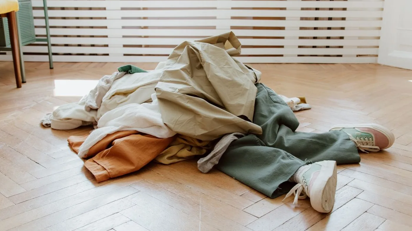 Heap of folded and crumpled clothing piled on a herringbone parquet floor beside a pair of trainers