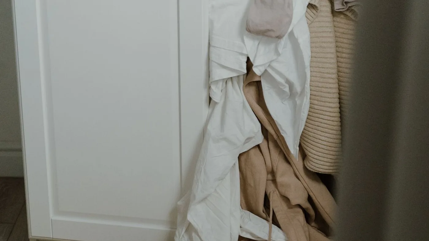 White fitted wardrobe with neutral shirts and knitwear spilling out of it onto a mustard rug