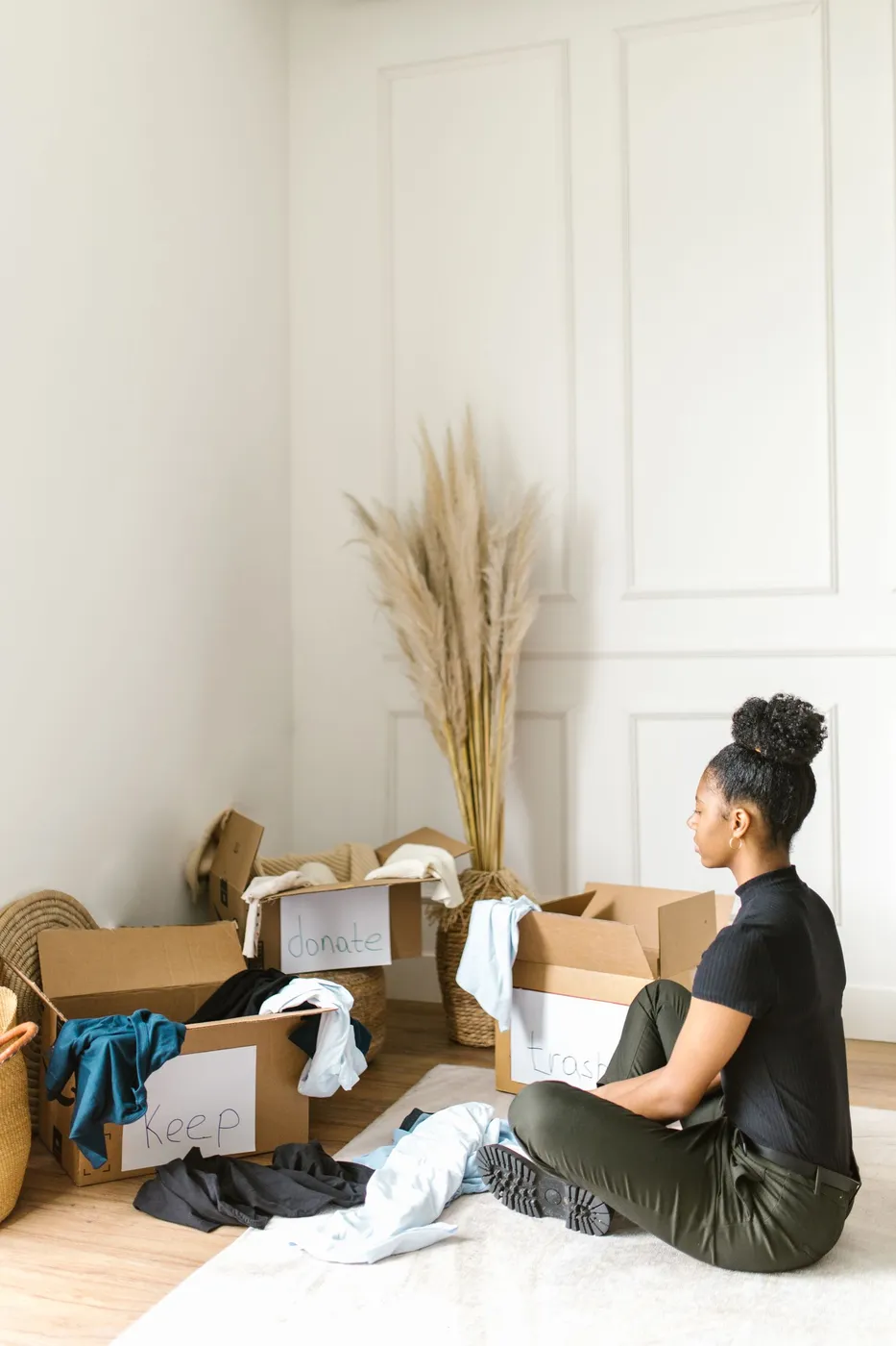 Woman sitting on a rug sorting clothes into open cardboard boxes hand-labelled keep, donate and trash