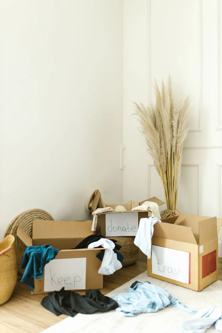 Three open cardboard boxes labelled keep, donate and trash with clothes spilling over their sides on a wooden floor