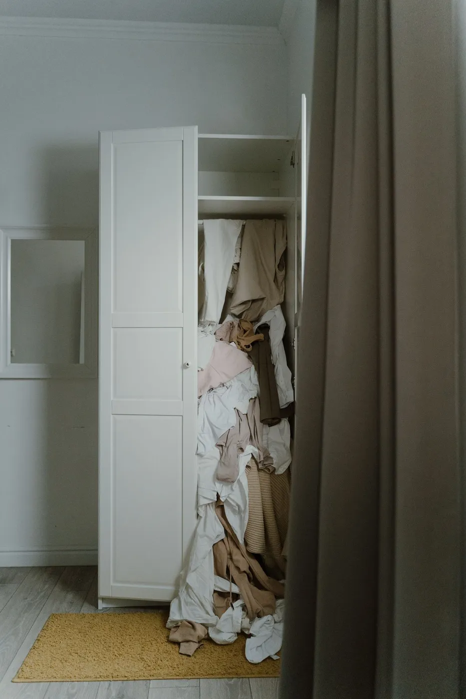 Open white wardrobe with neutral-toned clothes and bedding spilling out onto a yellow rug