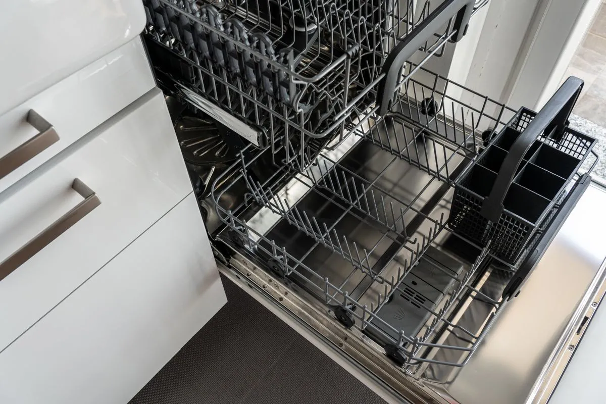 Built-in dishwasher with its door ajar revealing wire racks, set into white kitchen cabinetry beside a central island