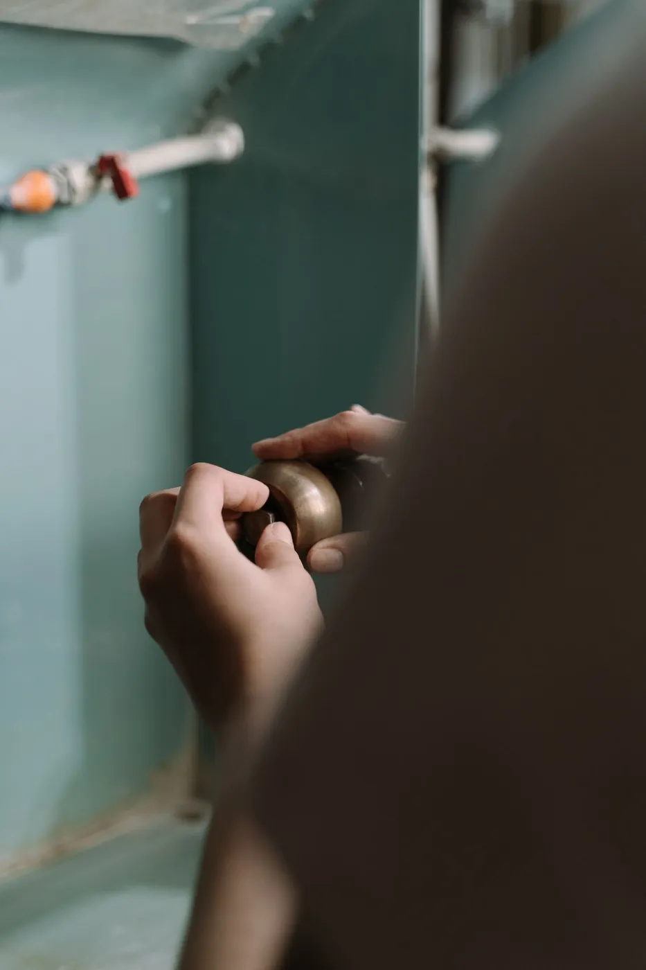 Close-up of a person's hands adjusting a copper pipe fitting inside a teal-painted kitchen cabinet, disconnecting a water supply line