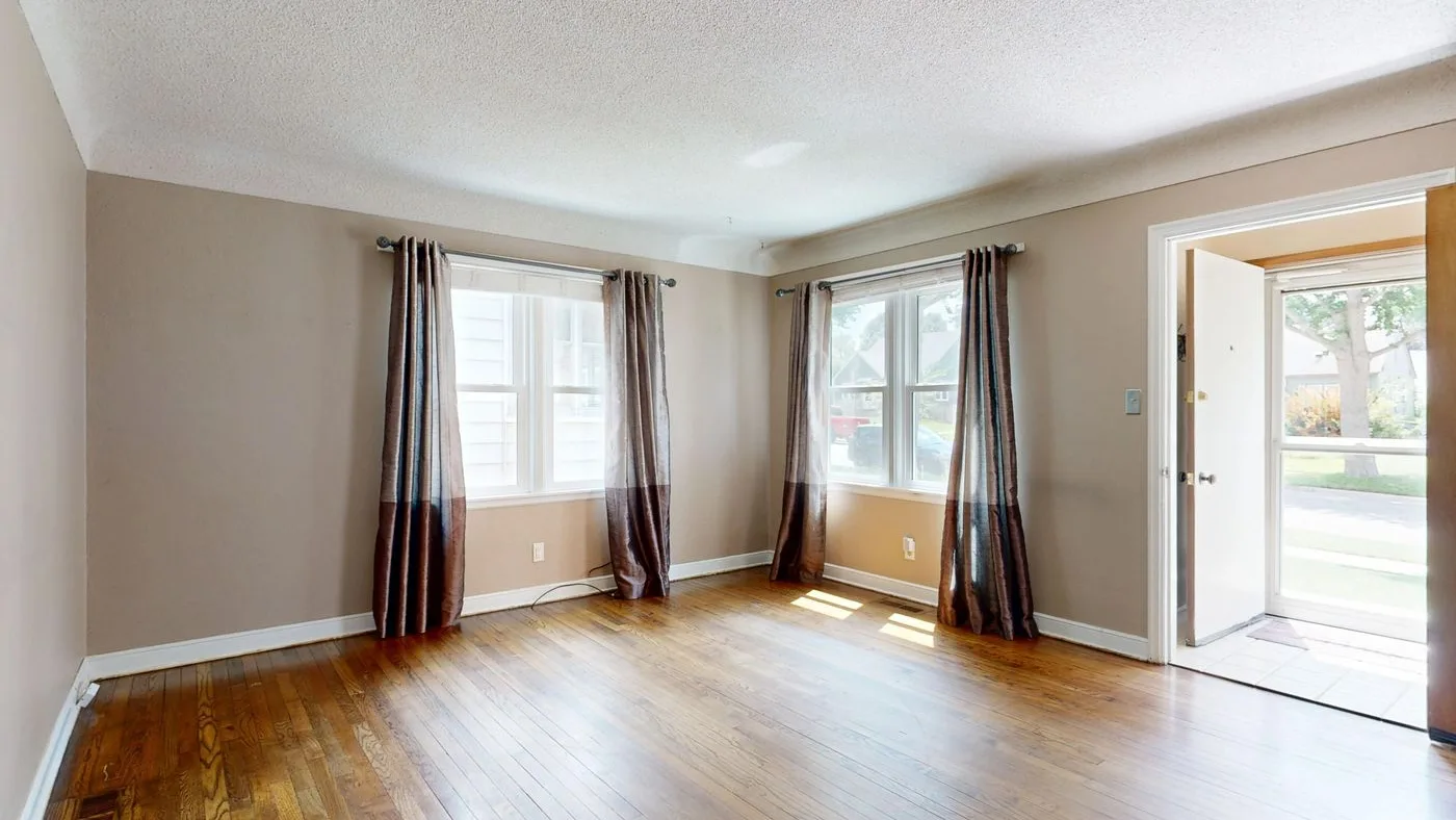 Emptied living room with bare wooden flooring, two curtained windows and an open front door showing a garden beyond