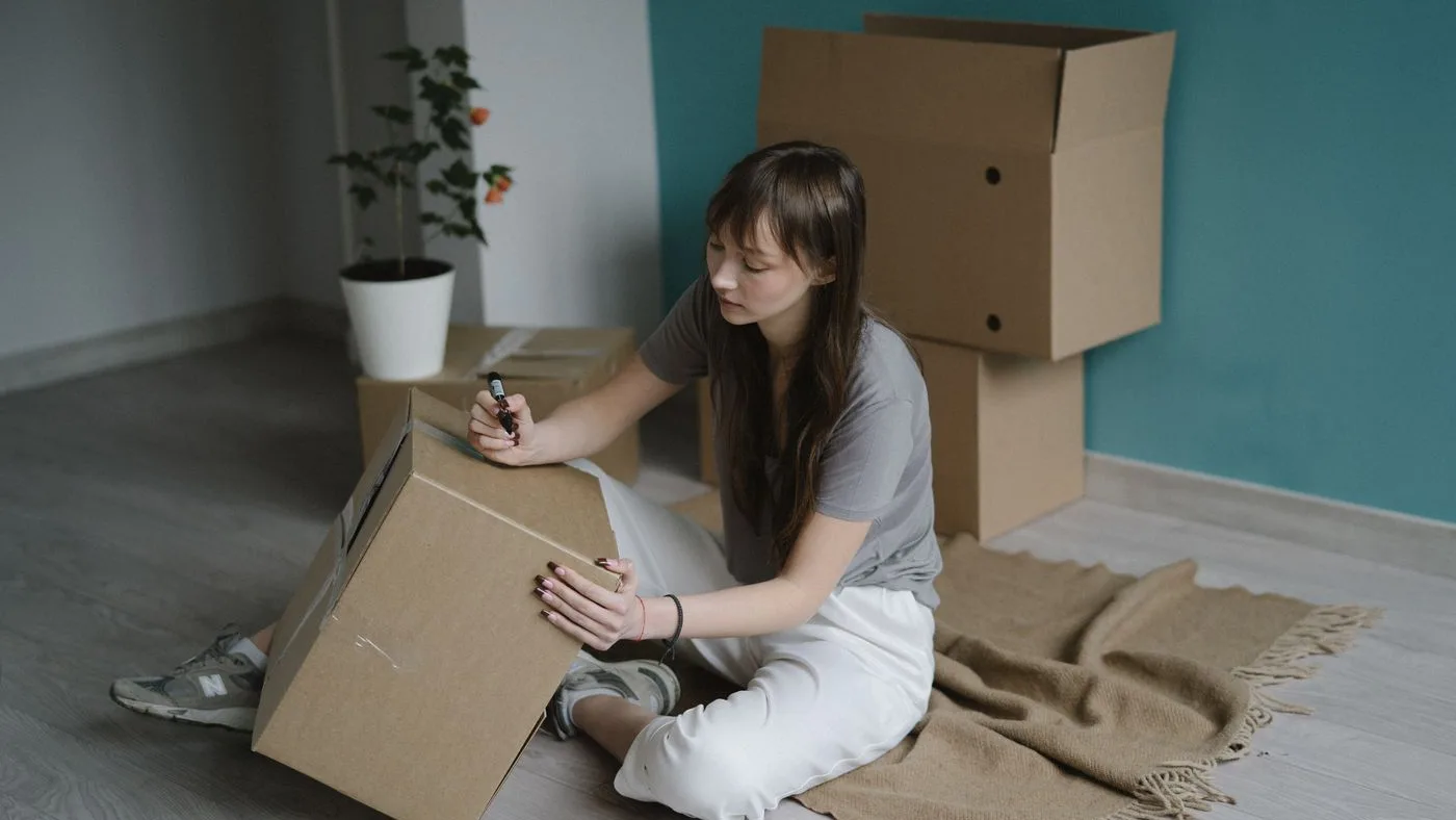 Person sitting on a wooden floor writing on a sealed cardboard box with a marker, with more boxes stacked against a teal wall