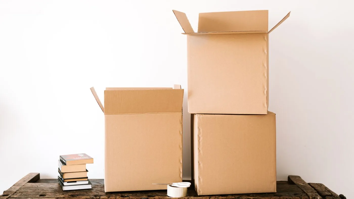 Two open cardboard boxes and a stack of hardback books with a roll of tape on a rustic wooden bench against a white wall