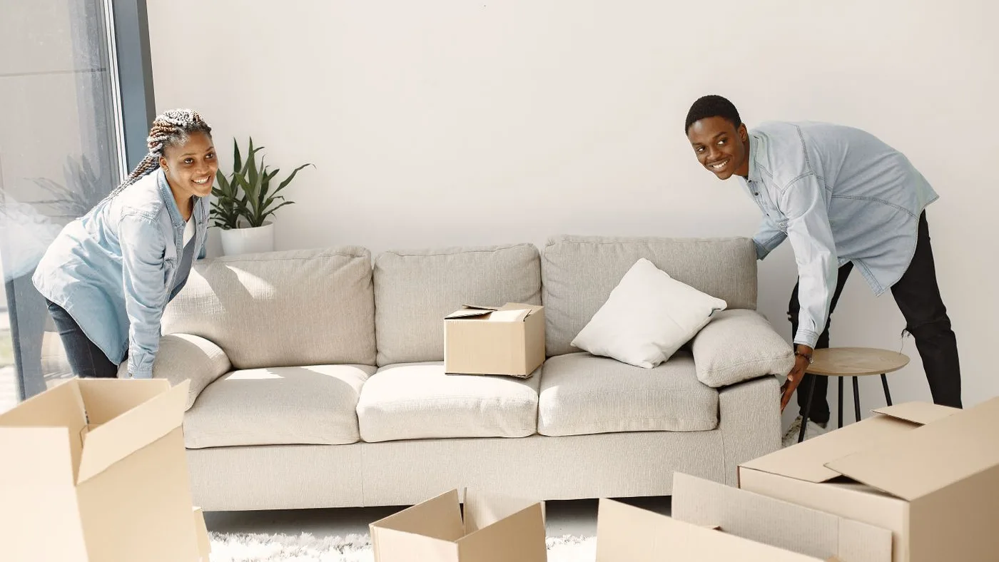 Man and woman at either end of a pale grey three-seater sofa, lifting it in a room with flattened cardboard cartons on the floor